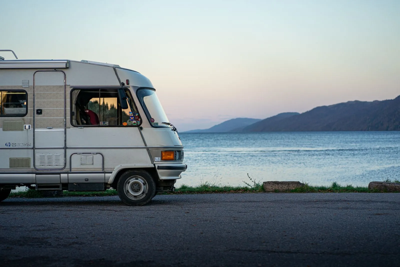 The front half of an older white motorhome parked on a lakeside road at dusk