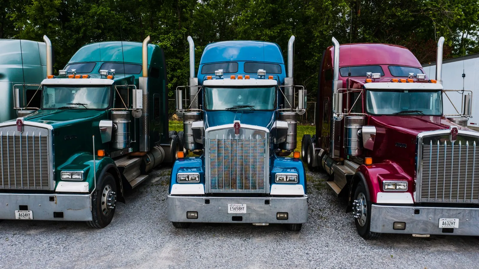 Three semi tractors, green, blue and maroon, parked side by side on a gravel lot